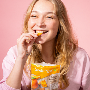 Eine Frau isst Cashew-Mango-Mix, lächelnd vor einem rosa Hintergrund.