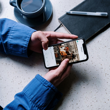 Hände halten ein Smartphone mit offenem Instagram. Daneben eine gefüllte Tasse und ein Notizbuch mit Stift.