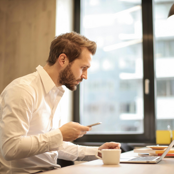 Ein Mann sitzt konzentriert mit Smartphone in einem modernen Büro vor einem Laptop.